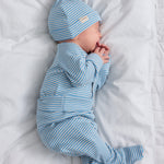 Baby iført blå- og hvidstribet cardigan og hue, liggende på hvidt tæppe