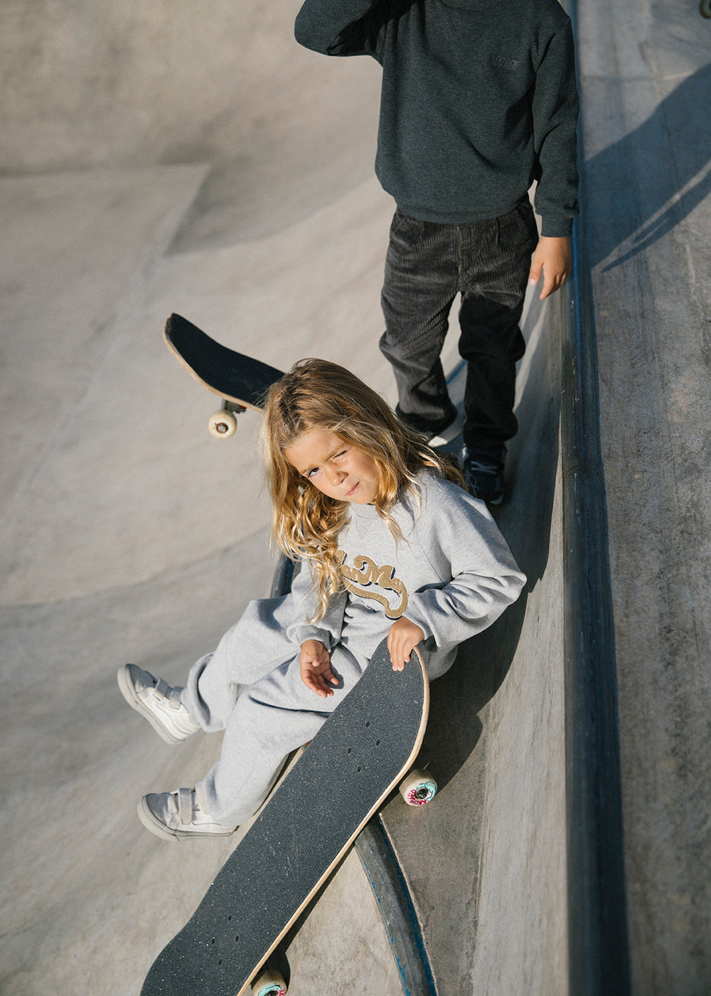 Børn med skateboard ved skatepark, iført grå sweatshirt og bukser