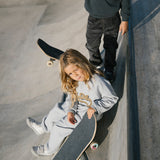 Børn med skateboard ved skatepark, iført grå sweatshirt og bukser