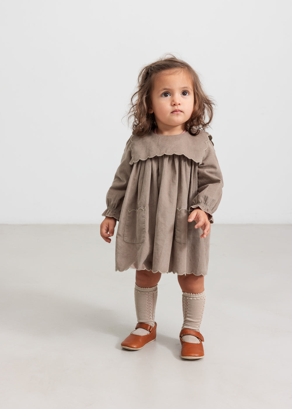 Pige iført beige babyfløjlskjole med lommer og lange ærmer