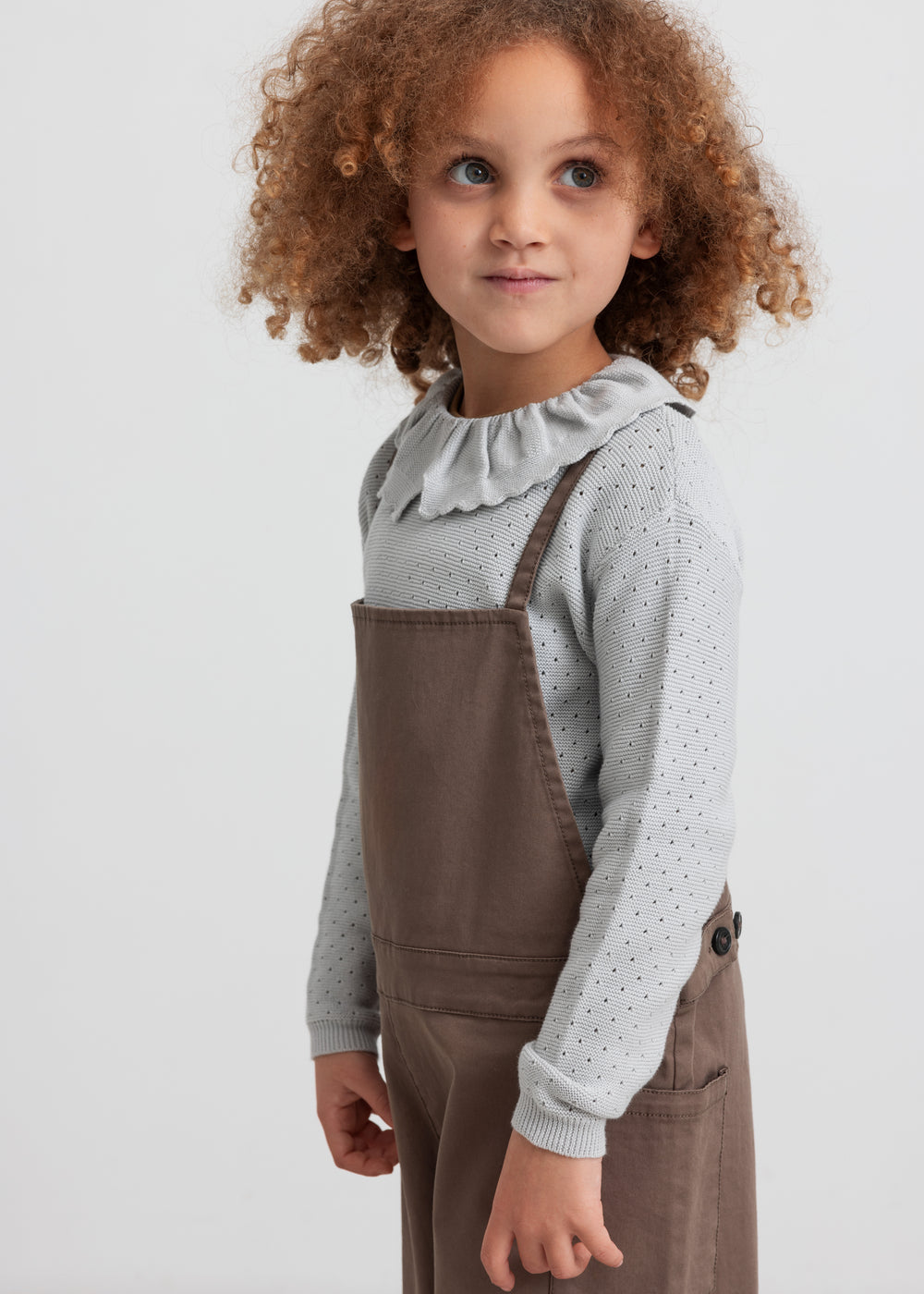 Pige iført brune selebukser og grå strikbluse set forfra