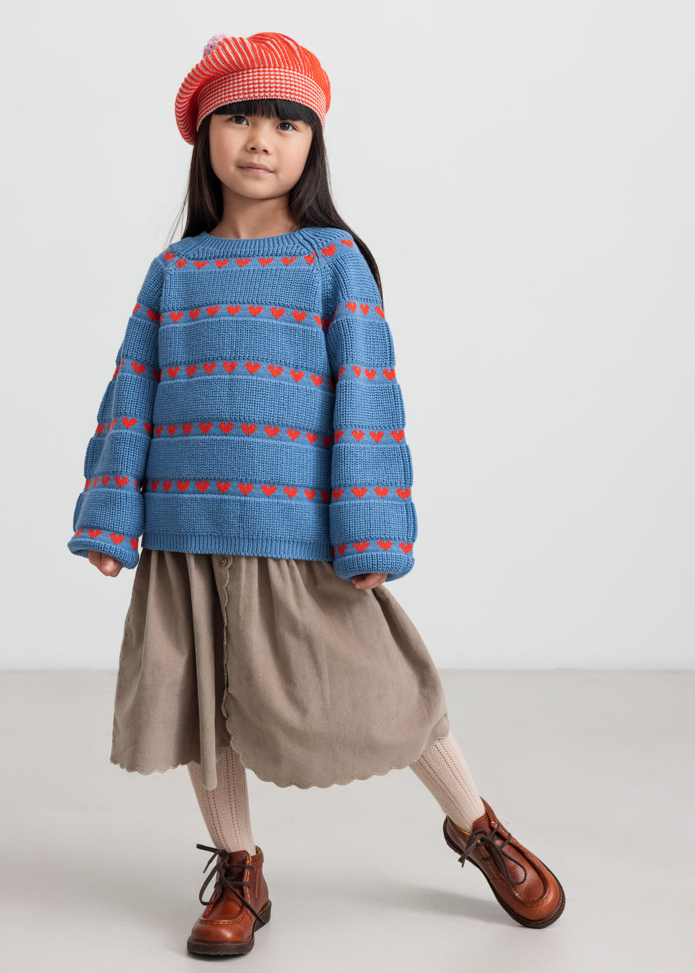 Børn iført blå striktrøje med røde hjerter, kombineret med brun nederdel og rød hue