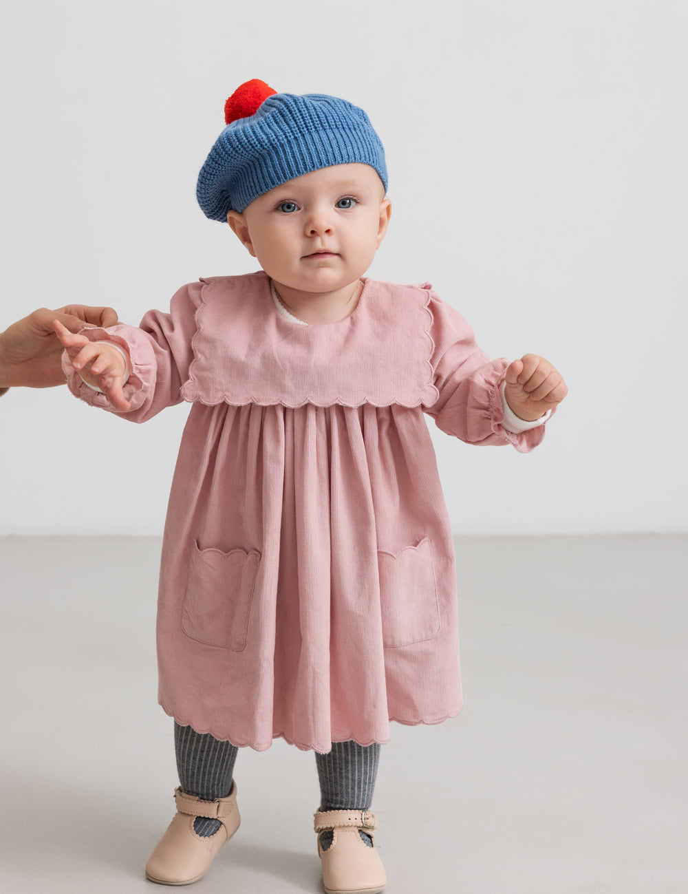 Baby i rosa kjole med blå strikket beret og rød kvast