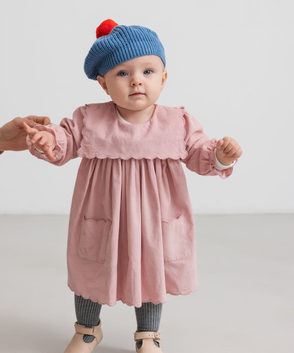 Baby iført lyserød babyfløjlskjole med lommer og blå beret med rød kvast