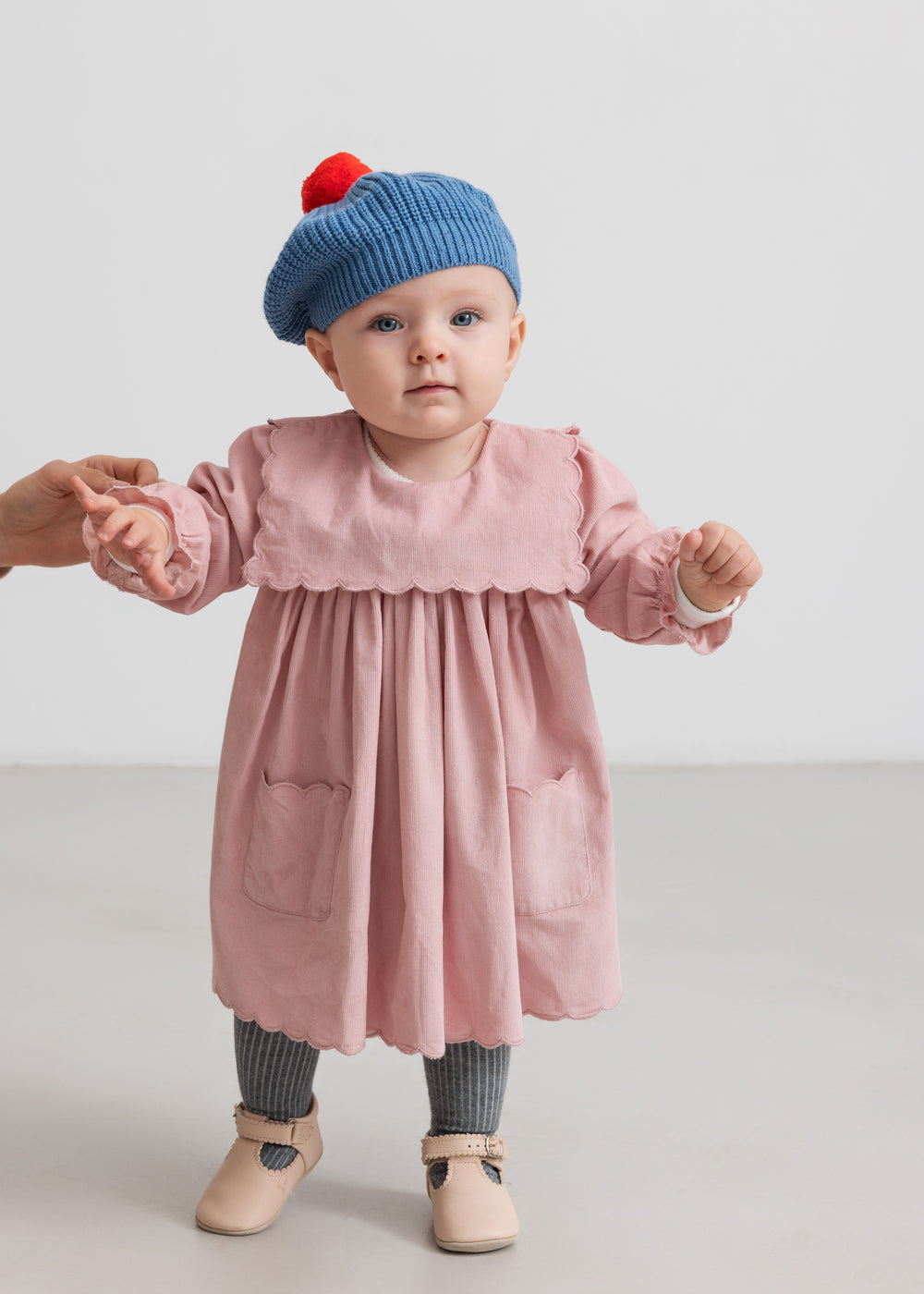 Baby iført lyserød babyfløjlskjole med lommer og blå beret med rød kvast