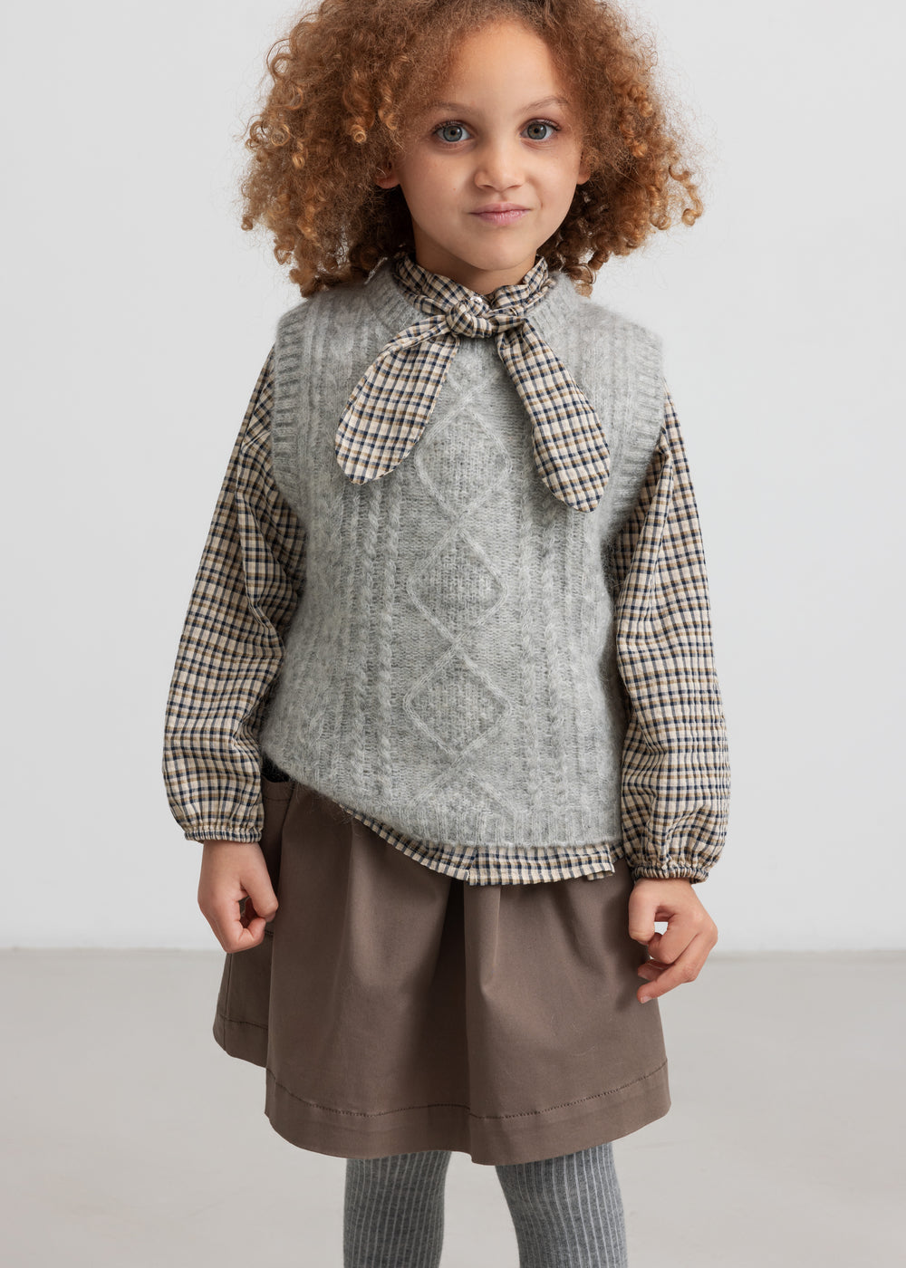 Pige iført brun ternet bluse under grå strikvest