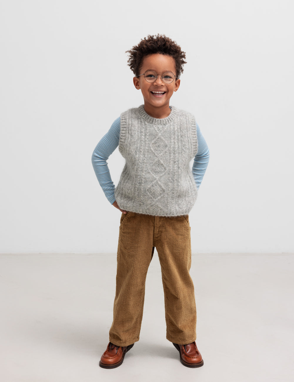 Dreng iført en blå og hvid stribet bluse med en grå strikvest og brune bukser.