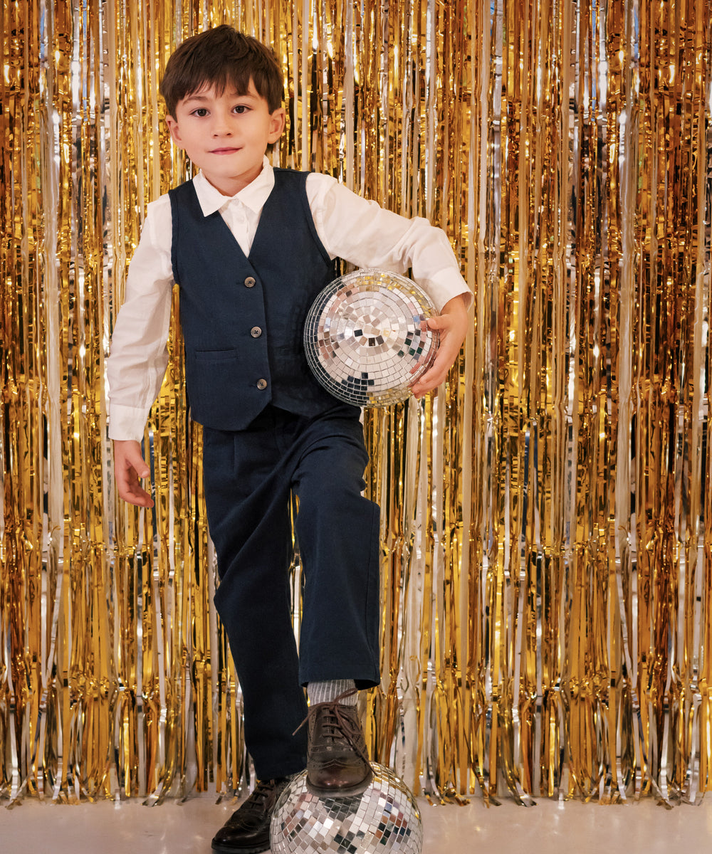 Dreng iført festtøj til børn mørkeblå bukser og hvid skjorte står foran gyldent festgardin med discokugle