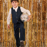 Dreng iført festtøj til børn mørkeblå bukser og hvid skjorte står foran gyldent festgardin med discokugle