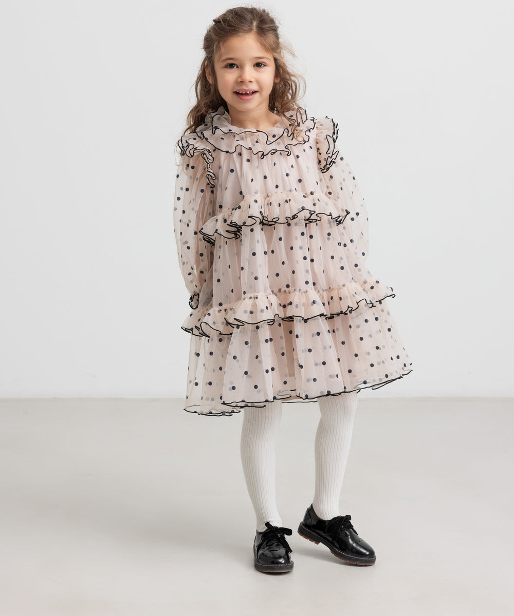 Pige iført lys beige julekjole i tyl med sorte prikker og hvide strømpebukser til børn