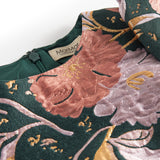 Nærbillede af mørkegrønt glansstof med blomsterprint og gyldne detaljer på festkjole til børn