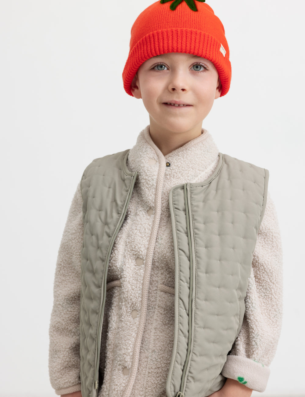 Barn iført grøn quiltet vest, teddy bluse og rød strikket hue