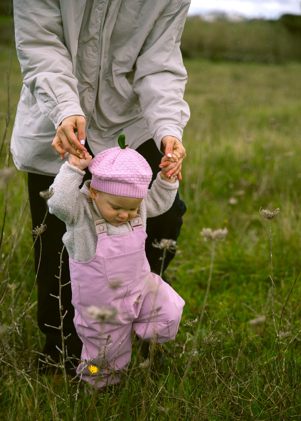 Baby iført lyserøde overtræksbukser og matchende hue gående støttet af voksen