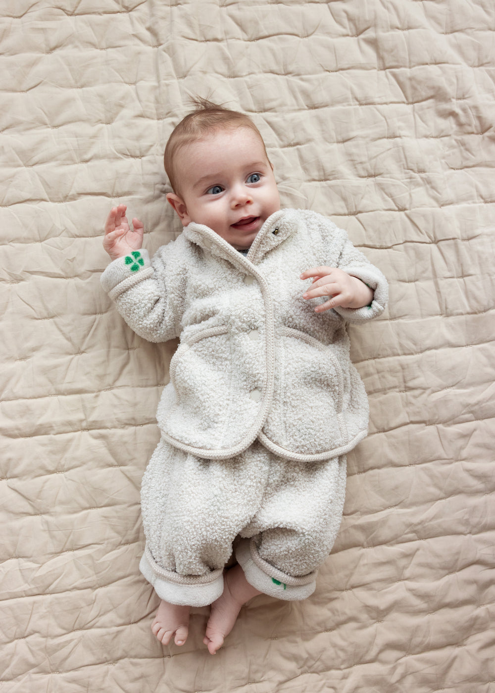 Baby iført beige fleecejakke og matchende bukser liggende på tæppe