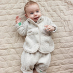 Baby iført beige fleecejakke og matchende bukser liggende på tæppe