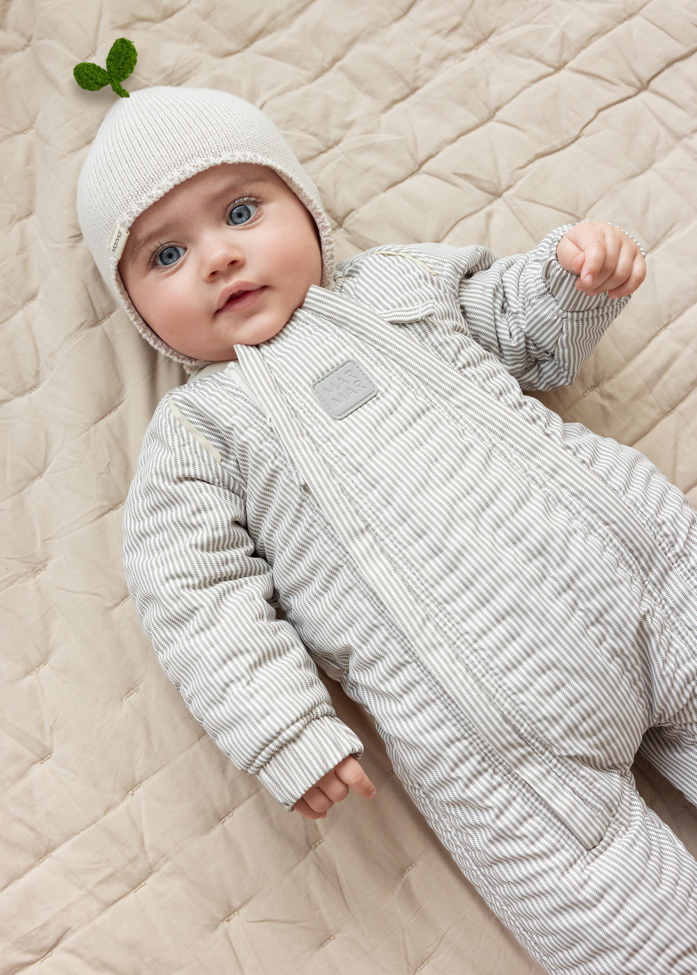 Baby iført brunstribet termodragt og beige hue liggende på tæppe