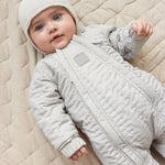 Baby iført brunstribet termodragt og beige hue liggende på tæppe