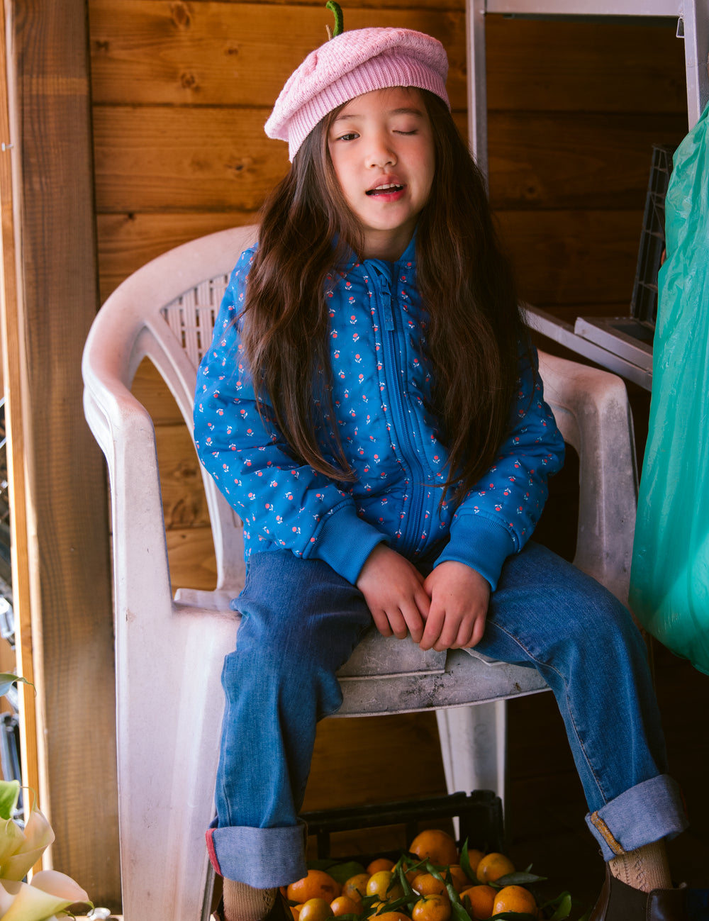 Barn i blå termo jakke med blomstret print og lyserød strikket hue siddende på stol