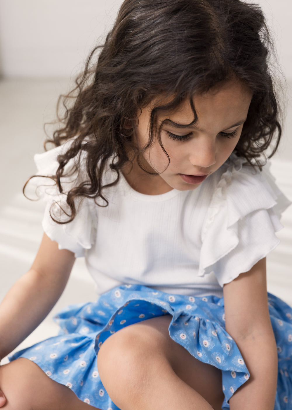 Pige iført kortærmet hvid pointelle t-shirt  med flæser på ærmerne og blå blomstret nederdel