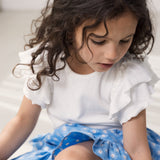 Pige iført kortærmet hvid pointelle t-shirt  med flæser på ærmerne og blå blomstret nederdel