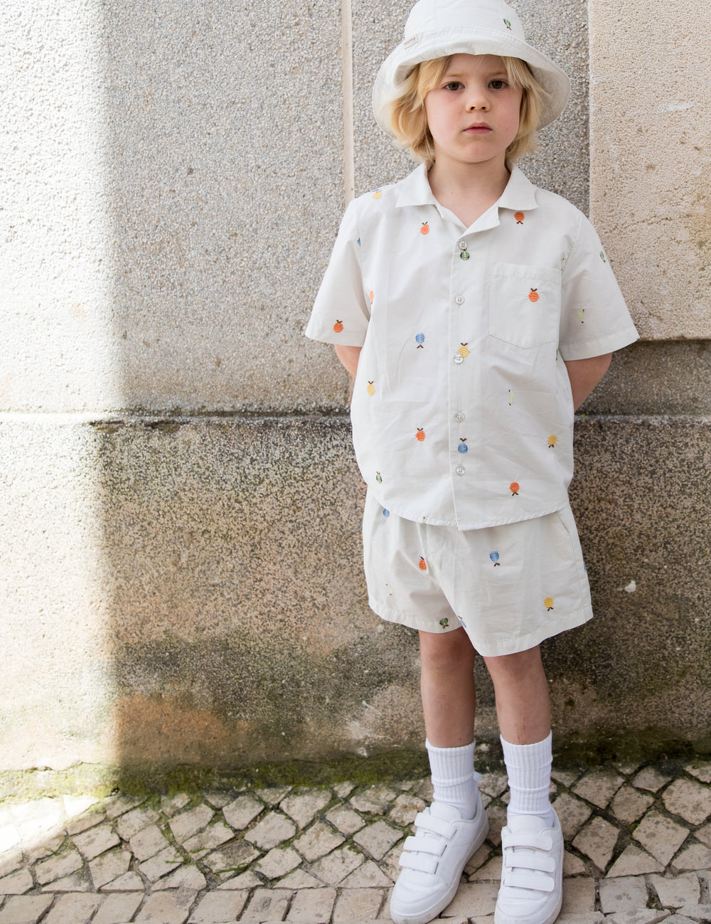 Barn iført lyst sæt med skjorte og shorts med frugtbroderi og solhat