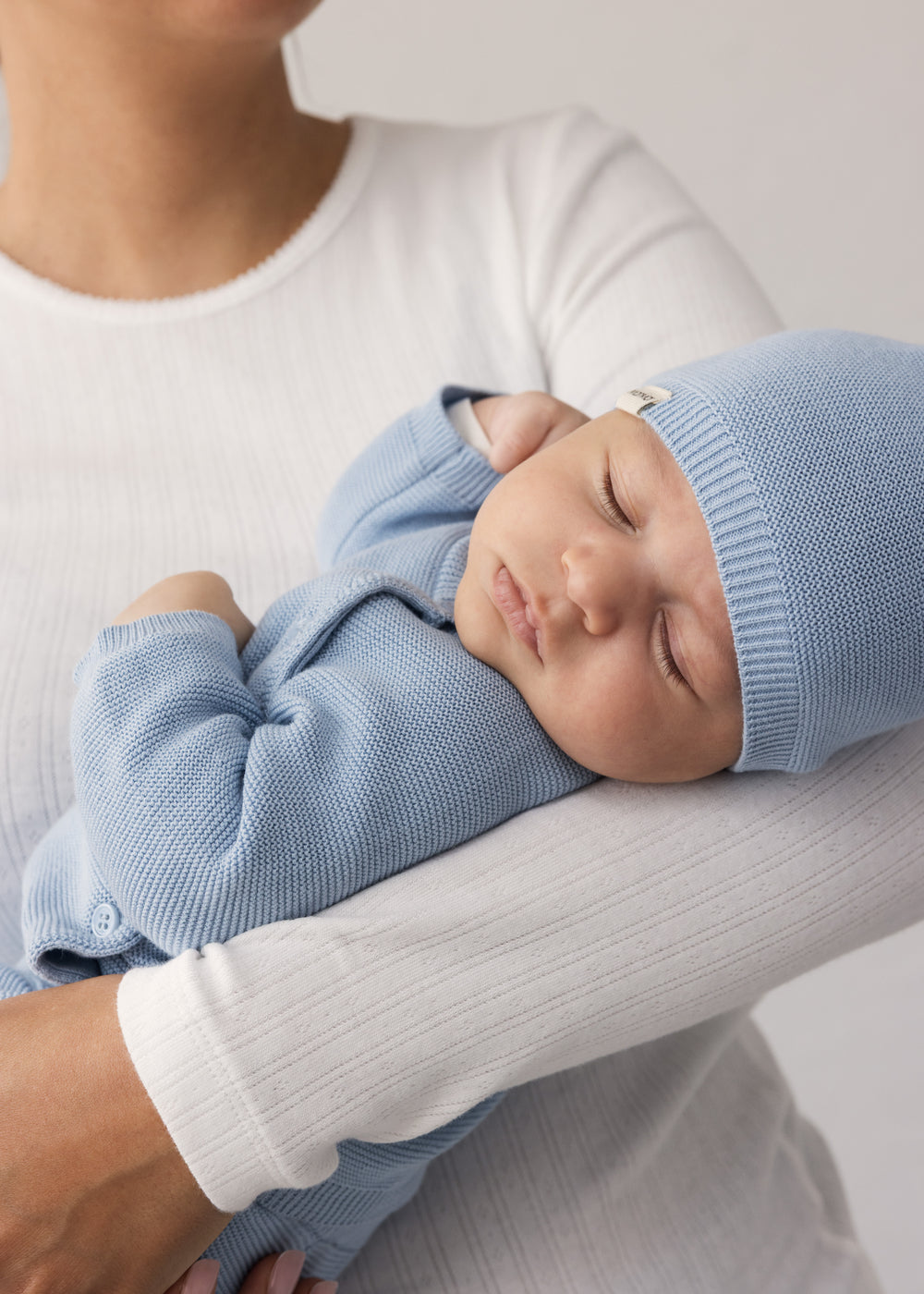 Kvinde i hvid t-shirt holder sovende baby iført blå hue og heldragt
