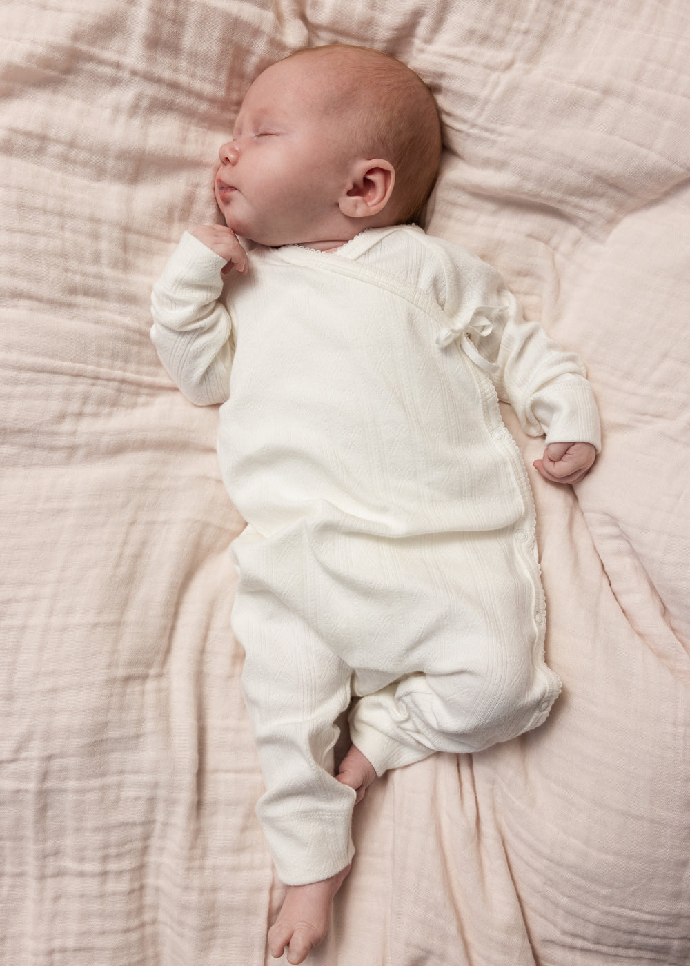 Hvid babyheldragt med knaplukning foran, båret af sovende spædbarn