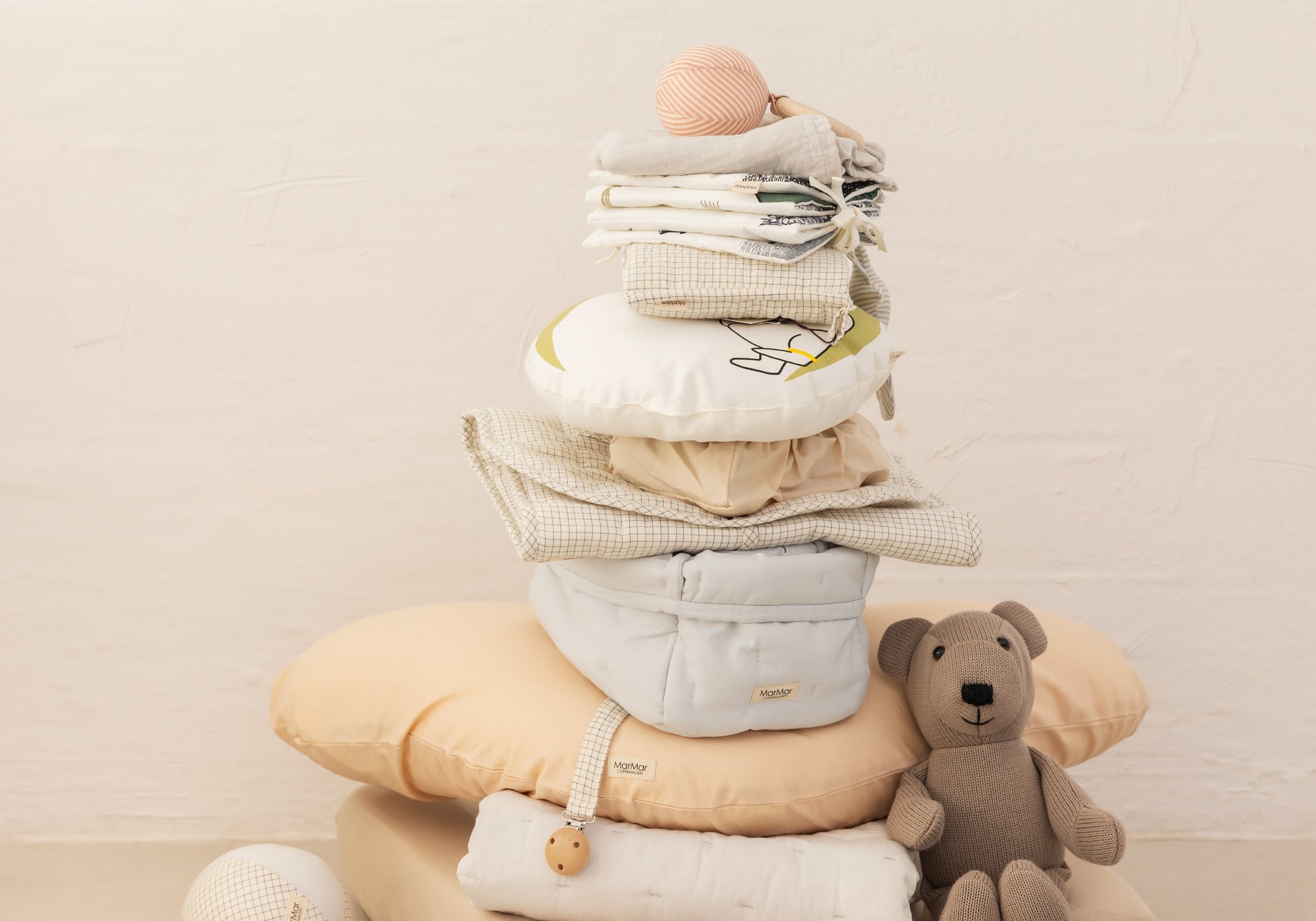 Stak af babytekstiler og puder i neutrale farver med strikket bamse ved siden af
