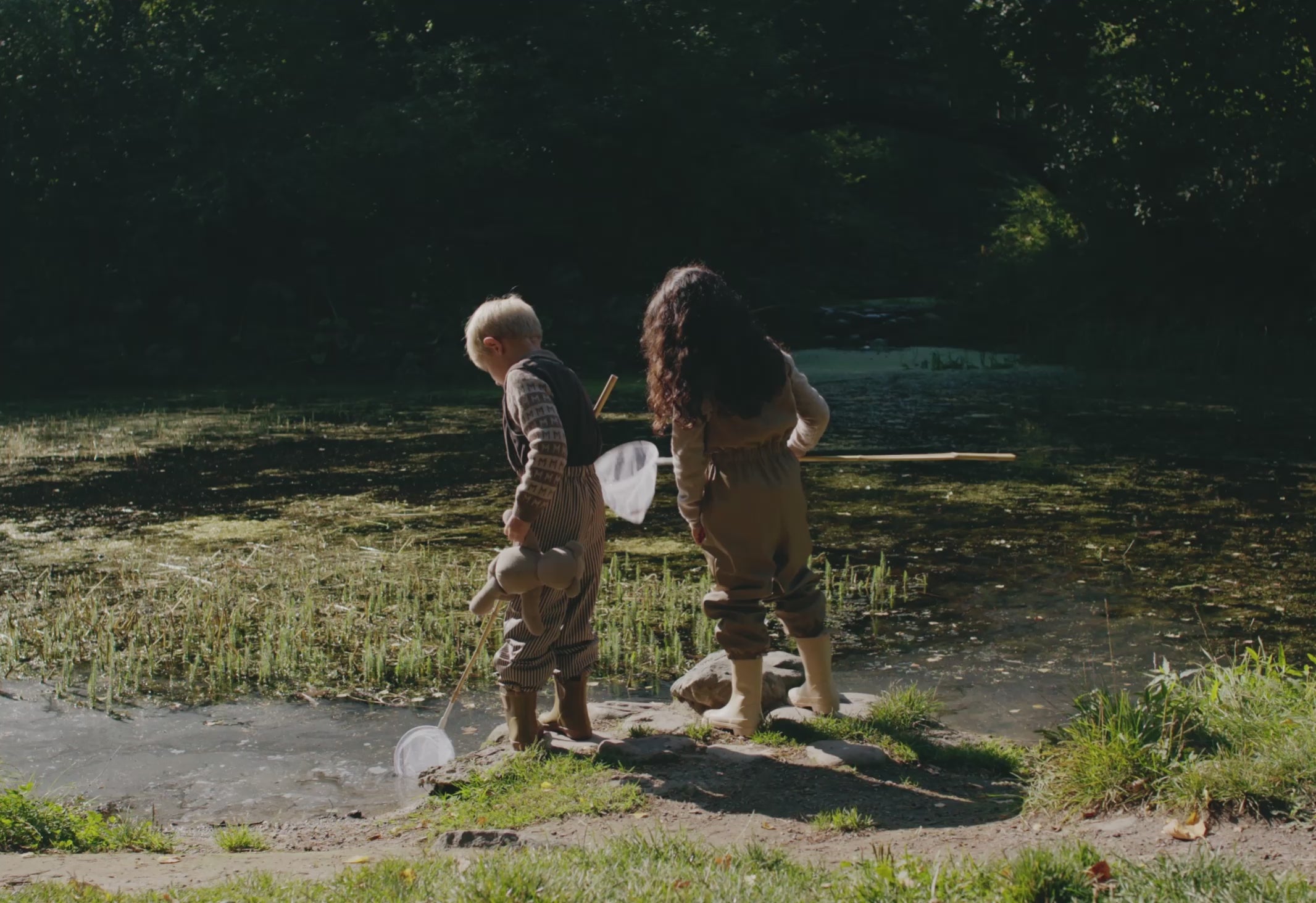 To børn leger udendørs ved vand i beige regntøj